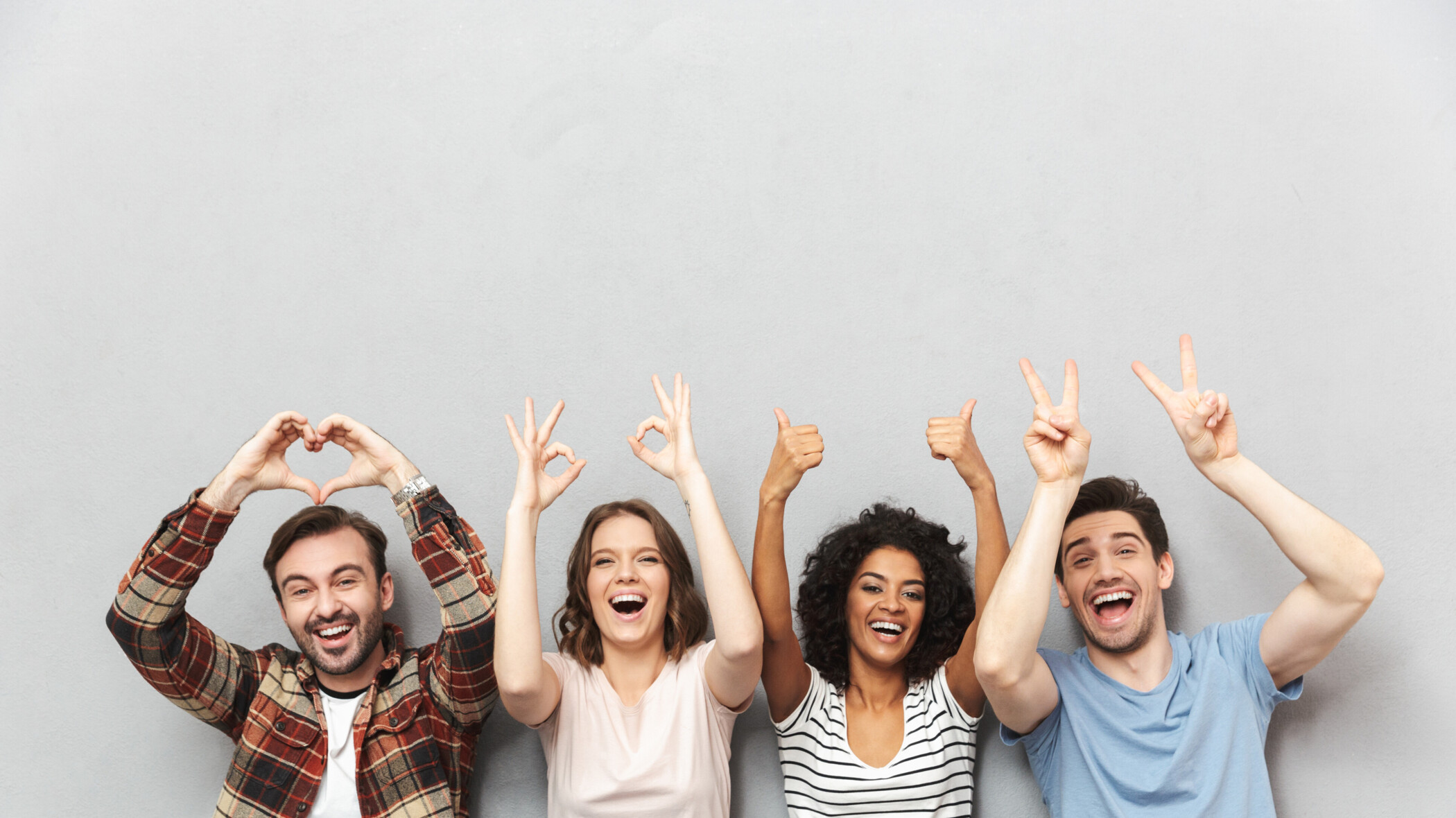 4 lachende junge Menschen, die die Arme in die Luft heben und mit den Fingern Herzen, Daumen hoch und das victory zeichen machen