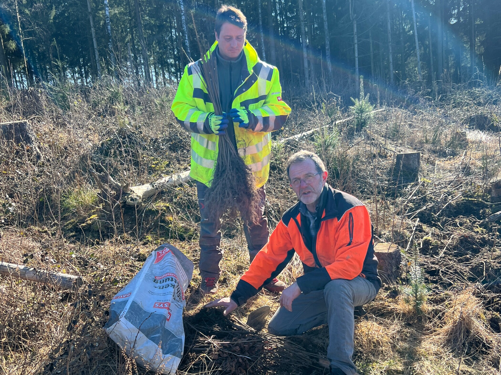 2 Baumpflanzer im Harz 2 Mitarbeiter der Stiftung Unternehmen Wald in Arbeitskleidung beim Pflanzen der Stecklinge, einer kniet auf dem Boden und schaut ins Bild, der andere steht und hält die kleinen Bäume in der Hand.